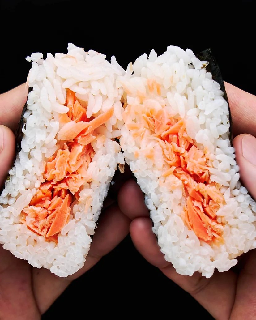 Two hands pulling a salmon onigiri apart to show flaked salted salmon inside the rice