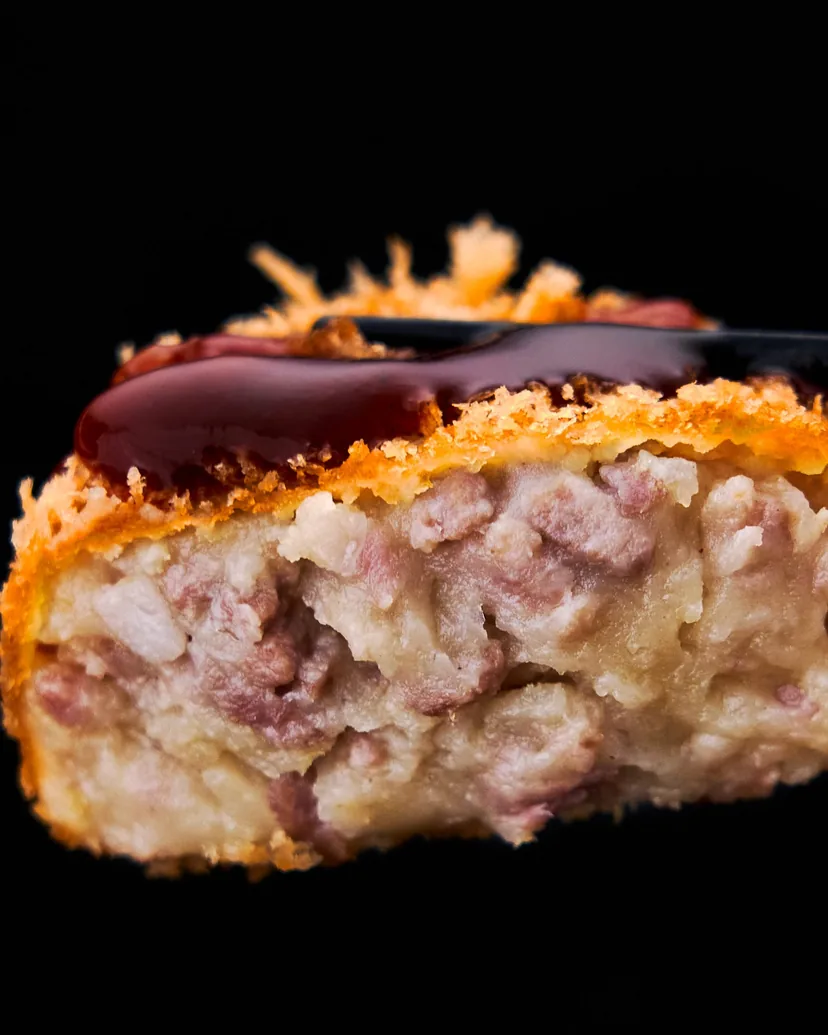 A potato and pork croquette cut open to show its filling, drizzled with sauce