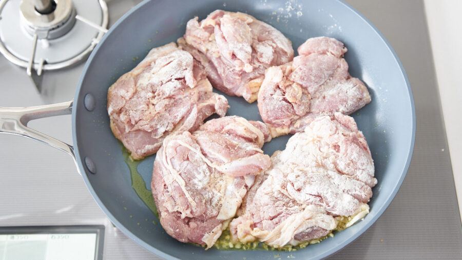 Chicken thighs browning in a frying pan with garlic for honey lemon chicken.