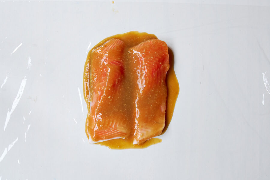 Salmon fillets coated evenly with miso paste on plastic-wrapped surface before wrapping.