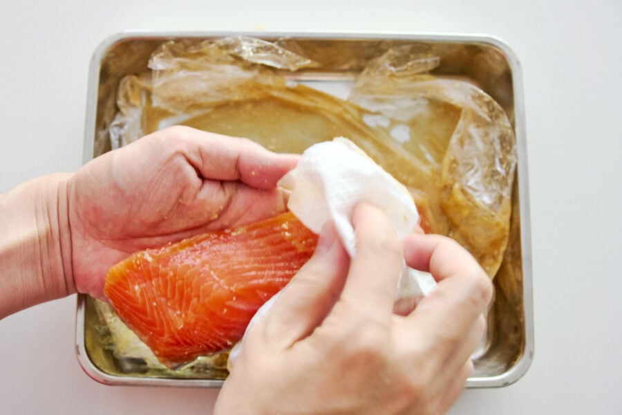 Hands wiping excess miso from a cured salmon fillet with a paper towel before cooking.