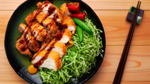 Crispy Chicken Katsu drizzled with katsu sauce on a green plate with shredded cabbage, tomatoes, and snap peas.