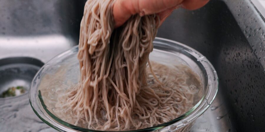 Rinsing boiled soba noodles under running water to remove excess starch from the surface of the noodles.