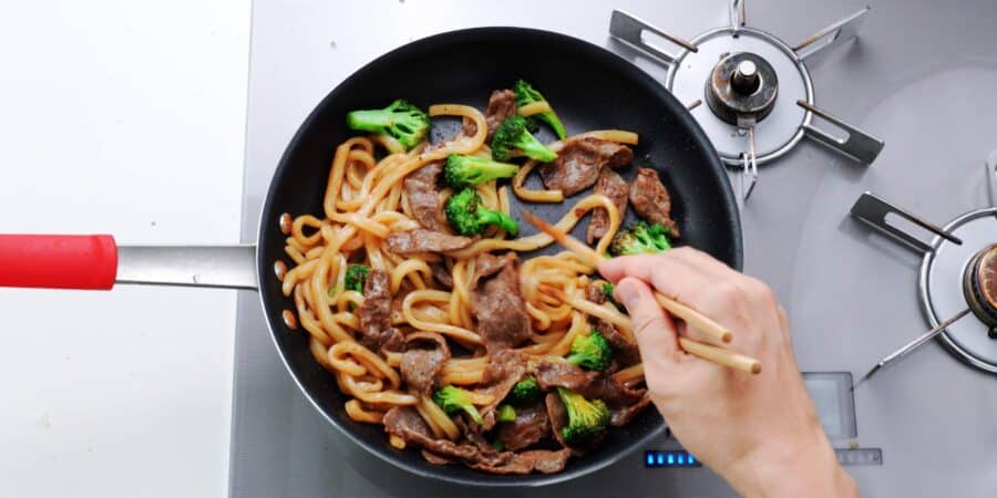 Stirring beef and broccoli yaki udon in a a large frying pan with chopsticks.