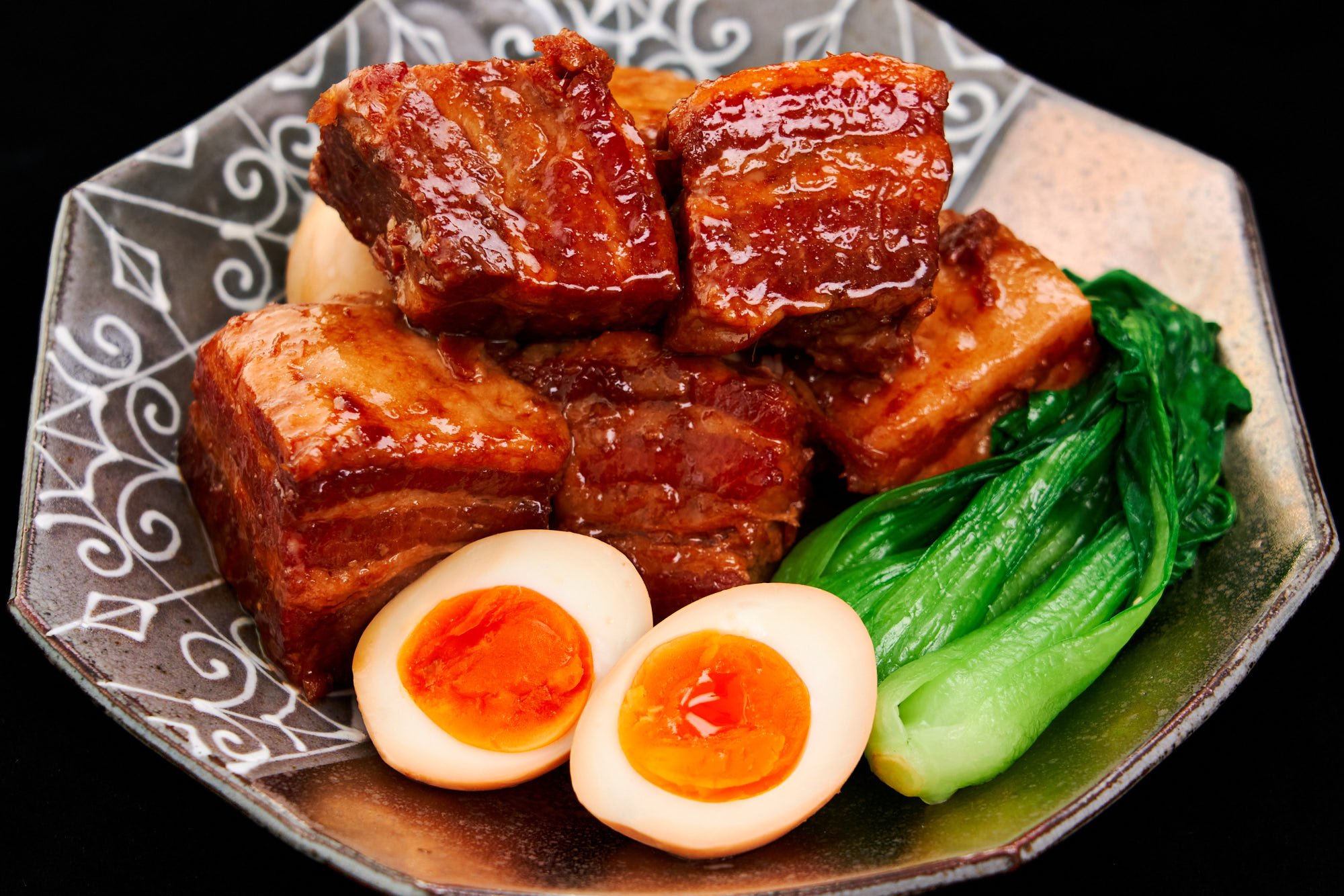 A close-up of luscious, slow-braised pork belly, shimmering with a savory-sweet glaze, nestled beside fresh bok choy and creamy soft-boiled eggs with golden centers.