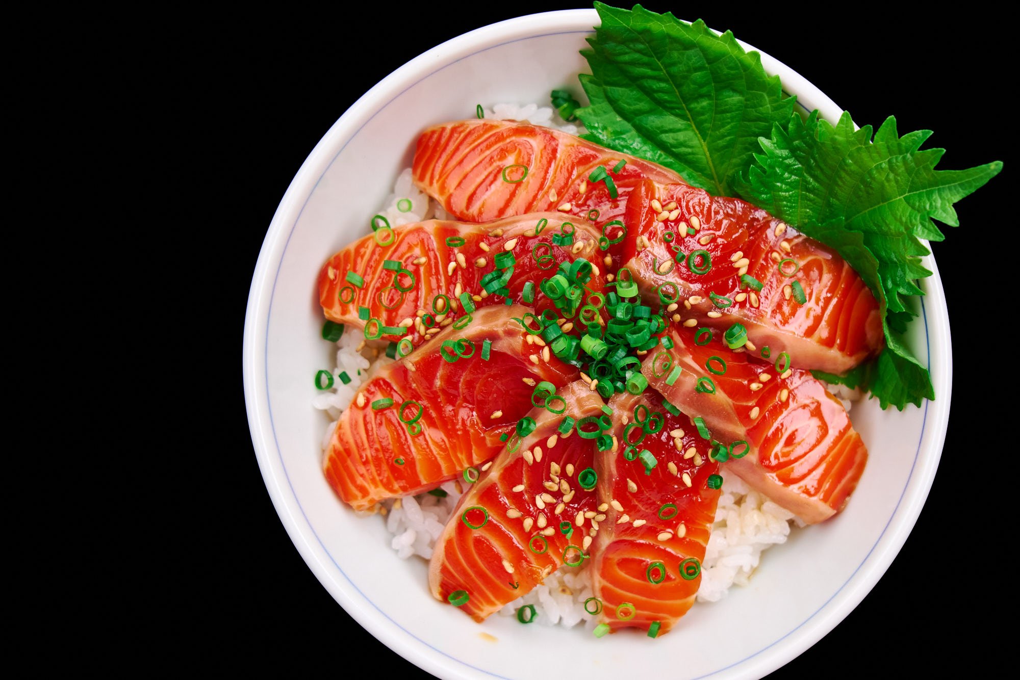 Silky slices of marinated salmon draped over fluffy rice, crowned with sesame seeds and scallions, framed by vivid green shiso.