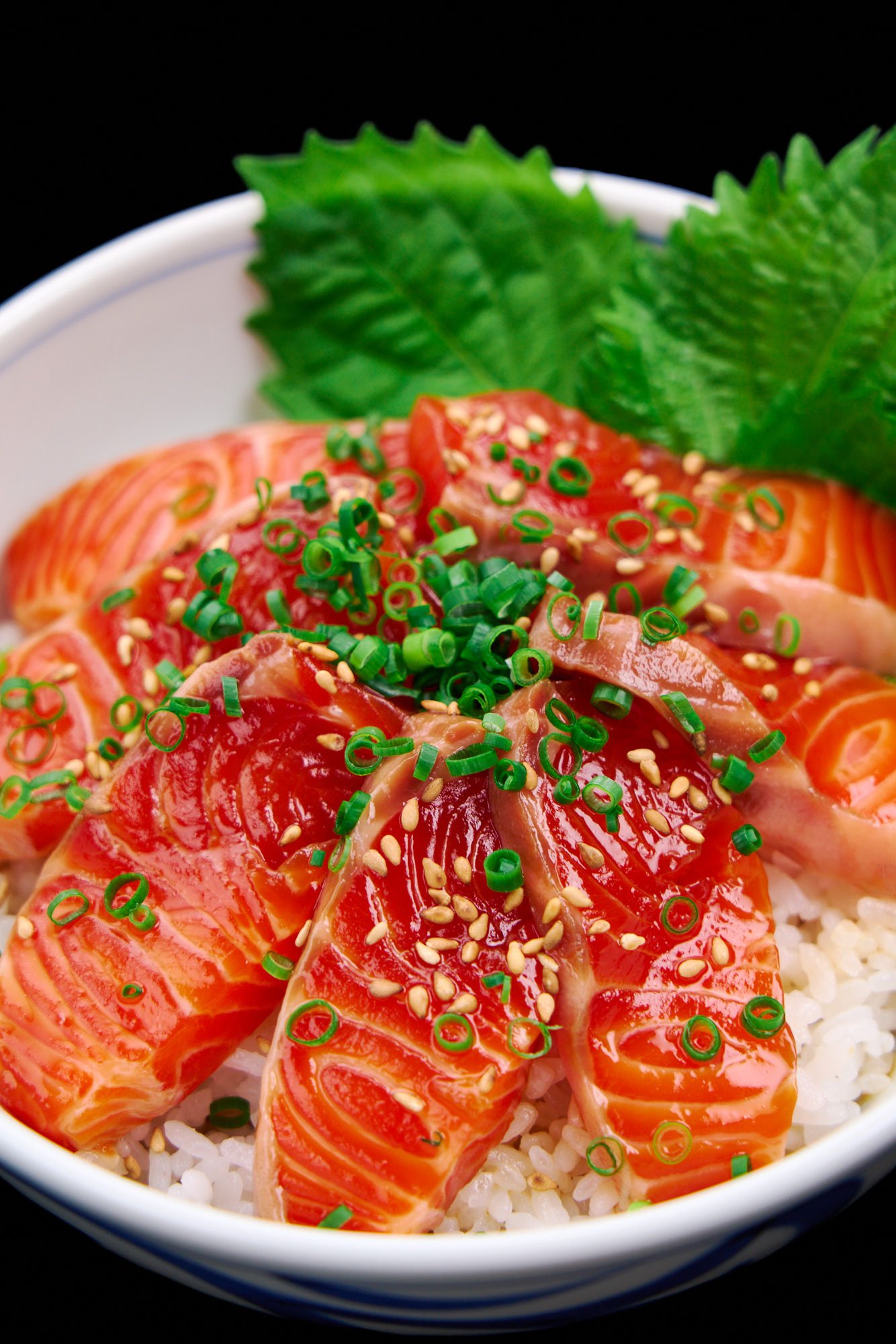 Close-up of a donburi bowl bursting with glossy salmon zuke, sprinkled with sesame and scallions, nestled against vibrant green shiso leaves.