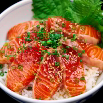 Close-up of a donburi bowl bursting with glossy salmon zuke, sprinkled with sesame and scallions, nestled against vibrant green shiso leaves.