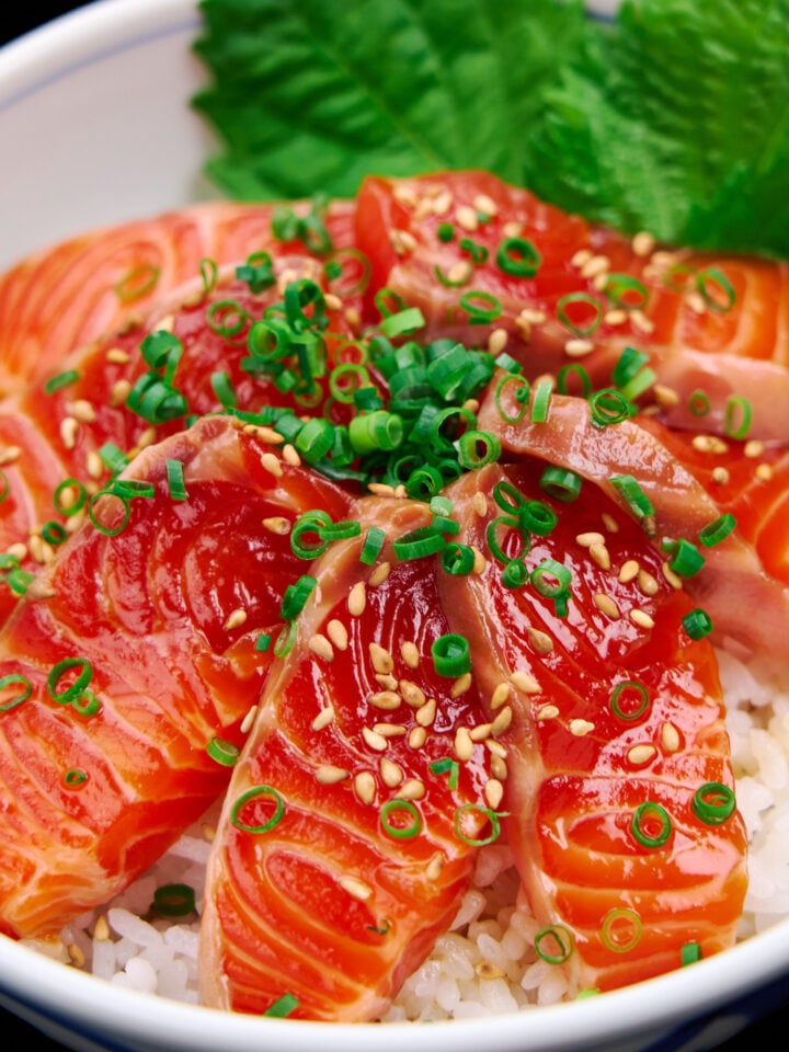 Close-up of a donburi bowl bursting with glossy salmon zuke, sprinkled with sesame and scallions, nestled against vibrant green shiso leaves.